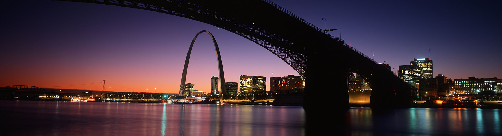 The Gateway Arch and St. Louis skyline at dusk, seen beneath a Mississippi River bridge